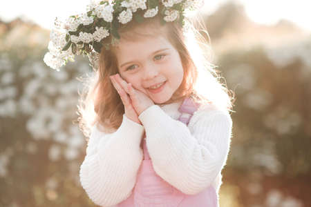 Smiling cute little child girl 3-4 year old wear floral wreath posing over white flowers at background. springtime. Childhood. Happy baby girl with flower headband over blooming bushes in garden.の写真素材