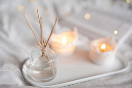 Home liquid fragrance in glass bottle and burning candles staying on white ceramic tray in bed close up. Cozy and hygge atmosphere.の写真素材