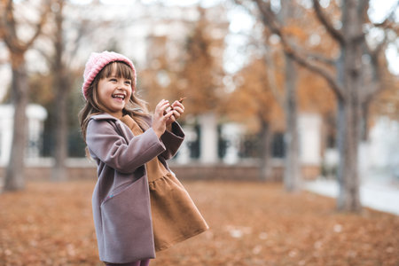 Laughing kid girl 4-5 year old playing with fallen leaves in park over nature background. Little toddler wear autumn jacket, dress and knit hat having fun. Happy cute babyの写真素材