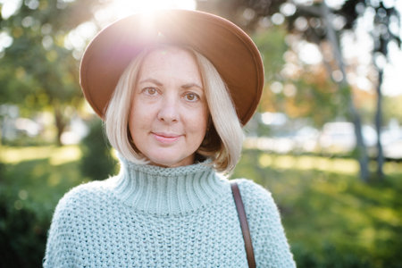 Smiling stylish blonde woman 50-55 year old wear hat and knit sweater walking in city street over...