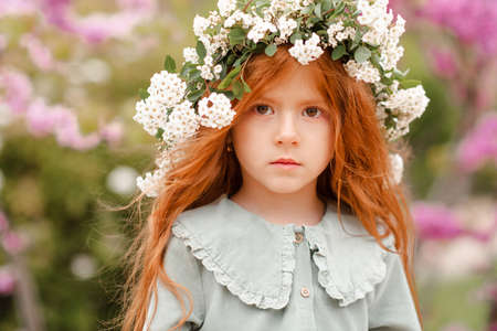 Cute little kid girl 3-4 year old with long curly red hair wear floral wreath and stylish rustic dress over nature background in garden outdoor. springtime. Calm pretty baby toddler in meadow.の写真素材