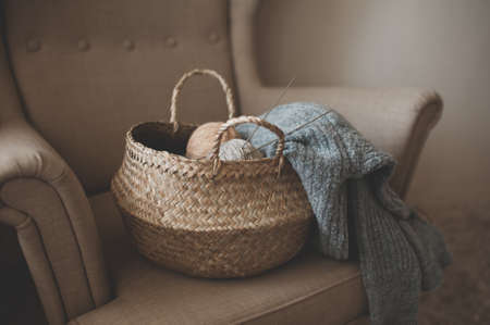 Making homemade knitted sweater at home. Straw basket with yarn wool skein with knitted fabric on armchair indoors. Having hobby. Cozy hygge atmosphere in the room.の写真素材