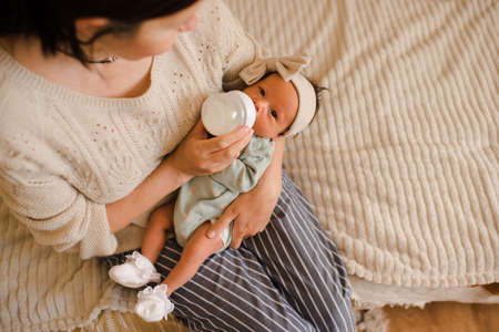 Young mother holding baby on hands in bed feeding with milk in plastic bottle top view close up. motherhood. Newborn child with woman wake up and eat in the morning at home. Healthy infant nutrition.の写真素材
