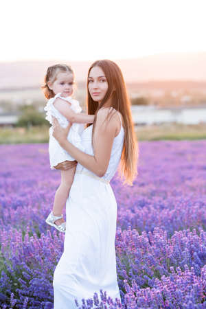 Beautiful young blonde woman holding baby girl stand in blooming lavender flower field outdoor together. motherhood. summer season.の写真素材