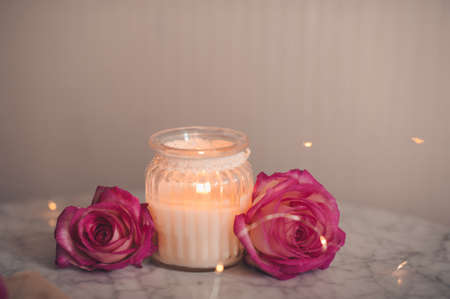 Burning scented candle in glass bottle with fresh rose flowers on marble table in room. Home cozy atmosphere.の写真素材