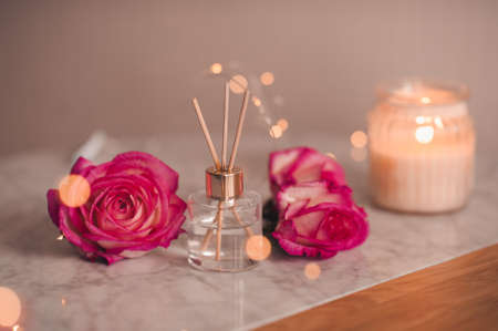 Liquid scented home fragrance in glass bottle and fresh rose flowers over burning candle and glow lights in room. Cozy atmosphere.の写真素材