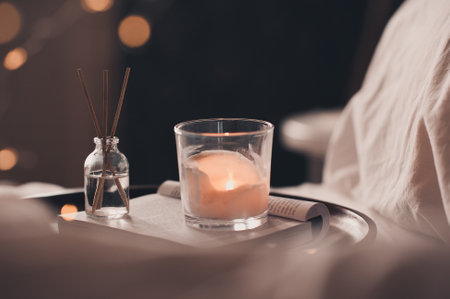 Home liquid fragrance on open paper book and burning scented candle on wooden tray and knit cloth sweater in bed close up. Cozy atmosphere.の写真素材