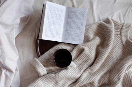 Open paper book with cup of coffee on wooden tray and knit cloth textile in bed close up top view....