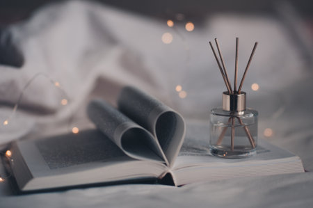 Paper open book with floded pages in heart shape in bed with bottle of liquid home scented perfume and bamboo sticks over glow lights. Cozy hygge atmosphere. aromatherapy.の写真素材