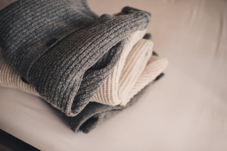 Stack of knitted wool textile sweaters clothes on white blanket in bed at home room close up. winter cozy season.の写真素材