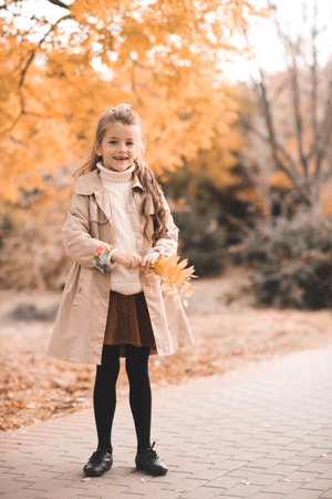 Smiling stylish child girl 5-6 year old wear knit sweater with yellow leaves over nature background outdoors. Happy little kid with blonde hairstyle. Childhood.の写真素材