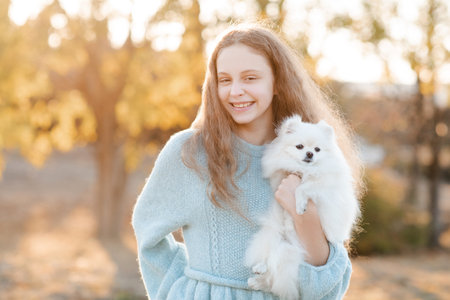 Cute smiling teenage girl 15-16 year old holding white fluffy spitz puppy dog together over autumn yellow nature background in park. Friendship.の写真素材