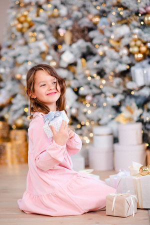 Smiling baby girl 5-6 year old open presents under Christmas tree over lights t background in room at home. winter holiday season. Childhood.の写真素材