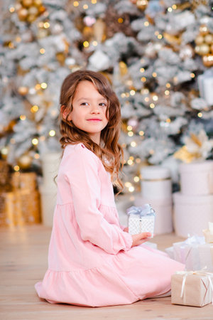 Smiling baby girl 5-6 year old open presents under Christmas tree over lights t background in room at home. winter holiday season. Childhood.の写真素材