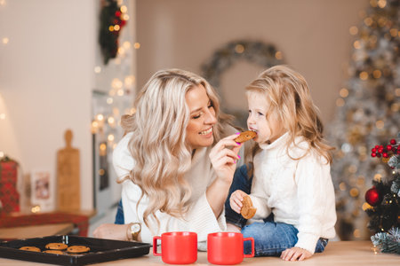 Happy mother with cute kid girl eating biscuits over Christmas tree and lights with decorations in home room. winter holiday season. celebration.の写真素材