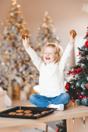 Funny child girl with tasty homemade biscuits over Christmas decoration and tree with lights at...