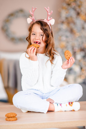 Little child girl 5-6 year old having fun eating homemade biscuits without front teeth sitting on kitchen table over Christmas tree lights wear deer headband closeup. winter holiday season. Childhoodの写真素材