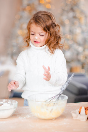 Smiling cute baby girl 3-4 year old cook Christmas cake over xmas tree lights in room at home kitchen. winter holiday season. Childhood.の写真素材