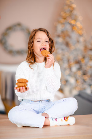 Cute funny child girl biting tasty biscuits sitting on kitchen table over Christmas tree lights in room at home close up. look at the camera. winter holiday season. Childhood.の写真素材