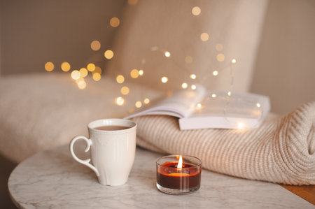 Coffee pot with cup of tea burning scented candle on marble table over Christmas lights in bedroom. Cozy home atmosphere.の写真素材