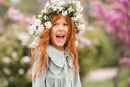 Cute laughing little kid girl 3-4 year old with long curly red hair wear floral wreath and stylish rustic dress over nature background in garden outdoor. springtime.の写真素材