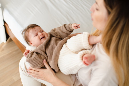 Sick newborn baby 4-5 months old with colic pain on mother hands in bedroom top view. Woman try calm her crying infant in bed. healthcare. motherhood.の写真素材