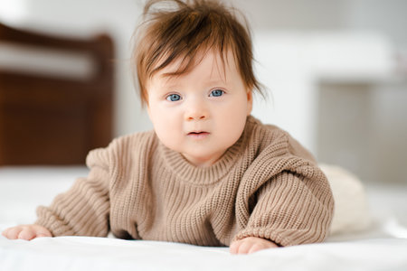 Cute funny newborn baby girl under 1 year old with blonde hair wake up wear beige knitted sweater crawling in bed in bedroom closeup. look at the camera. Childhood.の写真素材