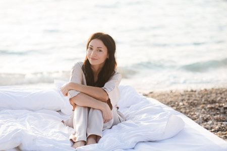 Smiling young woman wear silk pajamas sitting in bed with white duvet and pillows over sea shore outdoor. summer holiday seasonの写真素材