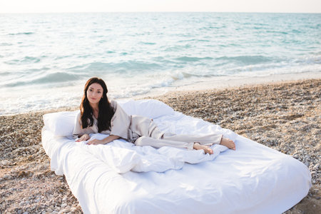 Smiling young woman wear silk pajamas sitting in bed with white duvet and pillows over sea shore...