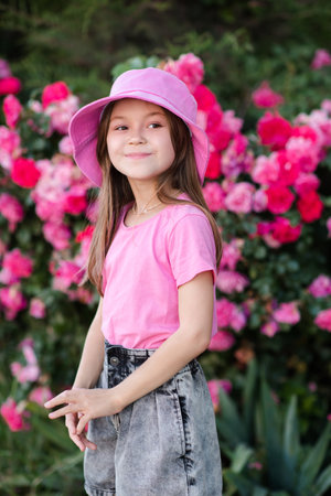 Smiling cute child girl 7-8 year old wear hat and pink shirt with denim shorts posing over blooming rose flowers in garden outdoor. summer season. Childhood.の写真素材