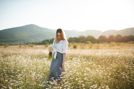 Beautiful attractive blonde woman pick up chamomile flowers walk in blooming meadow outdoor. spring season.の写真素材