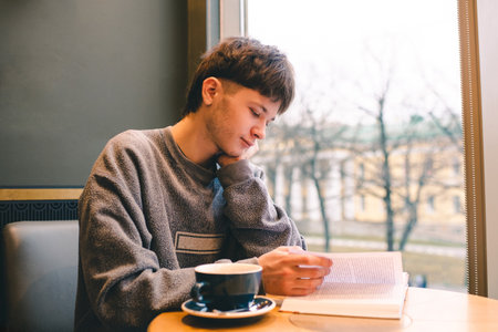 Teenage blond boy 18-19 year old reading book and drinking coffee in cafe alone.の写真素材