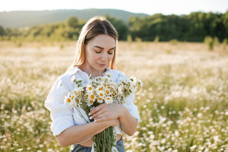 Beautiful attractive blonde woman 25-27 year old pick up chamomile flowers walk in blooming meadow outdoor. spring season.の写真素材