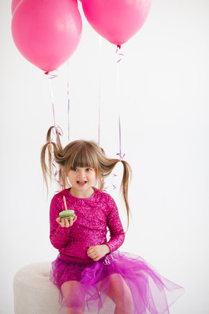 Cute happy child girl 4-5 year old wear princess dress posing with balloons eating cake with candle having birthday party over white in room. Childhood.の写真素材