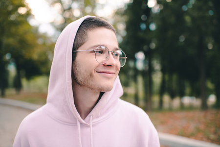 Smiling cute teenager boy 19-20 year old wear glasses and hood over nature background outdoor close up. Happy male kid student.の写真素材