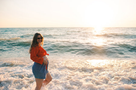 Pregnant happy woman walking at sea shore over sun light wearing casual prenatal clothes at beach. motherhood. summer holiday season.の写真素材