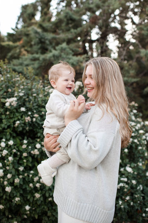 Happy smiling young mother holding baby 1 year old wearing stylish casual knitted clothes standing over blooming bushes with flowers in park outdoor. motherhood. maternity.の写真素材