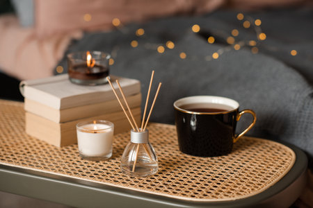 Home liquid fragrance in glass bottle with bamboo sticks with cup of black tea and scented candle on stack of paper book over glow Christmas light in bedroom closeup. aromatherapy. winter holiday.の写真素材