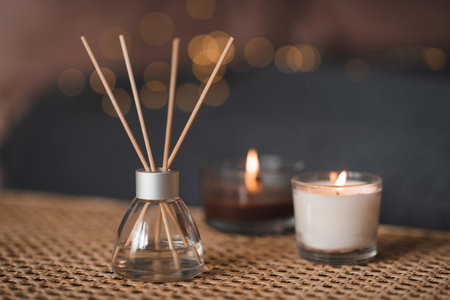 Liquid home fragrance in glass bottle and bamboo sticks with scented candle on rattan table over glow light in bedroom closeup. Cozy hygge atmosphere. Coziness.の写真素材