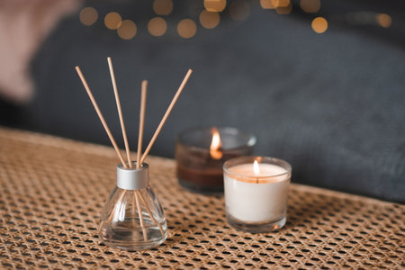 Liquid home fragrance in glass bottle and bamboo sticks with scented candle on rattan table over glow light in bedroom closeup. Cozy hygge atmosphere. Coziness.の写真素材