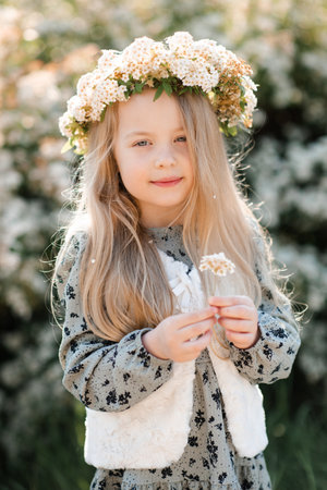 Smiling stylish child girl 4-5 year old wear floral wreath hairstyle and dress standing over flower at background outdoor. Summer season.の写真素材