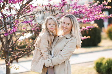 Happy young mother holding smiling cute kid girl 4-5 year old wear similar beige trench coats over blooming tree with pink flowers in park outdoor. Motherhood. Springtime.の写真素材