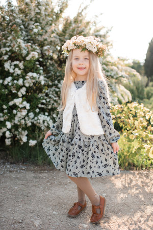 Smiling stylish child girl 4-5 year old wear floral wreath hairstyle and dress standing over flower at background outdoor. Look at camera. Summer season.の写真素材