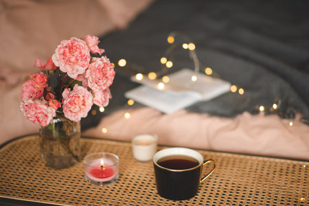 Cup of tea with scented candle and bouquet of rose flowers on rattan table over bed with glow lights closeup. Hygge cozy atmosphere.の写真素材