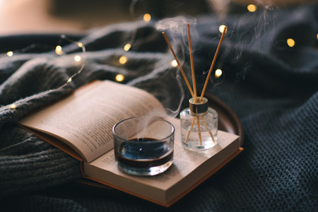 Scented candle with aroma diffuser on paper book over glow lights in bed close up. Aromatherapy. Cozy home atmosphere.の写真素材
