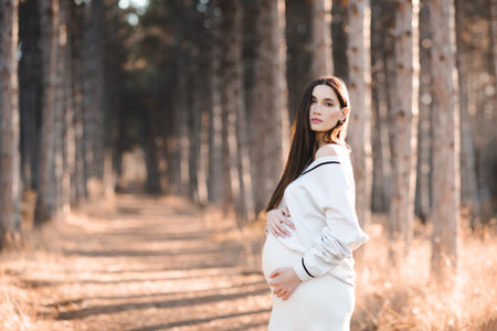 Stylish pregnant woman 25-30 year old wear knitted sweater holding her belly in woods outdoor. Maternity lifestyle. Autumn season. Looking at camera.の写真素材