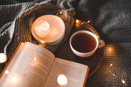 Cup of black tea with burning candle with open paper book on tray in bed over glowing Christmas lights close up. Cozy home atmosphere. Winter holiday season.の写真素材
