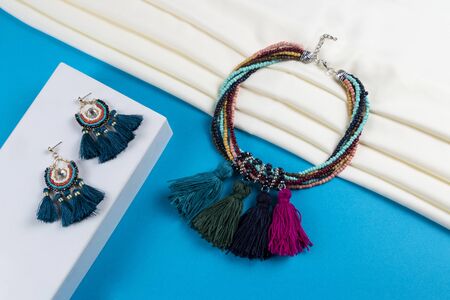 set of tassel earrings and multi layer multi color chunky seed beads with colorful tassels and crystal beads necklace boho style on white scarf and blue paper backgroundの写真素材