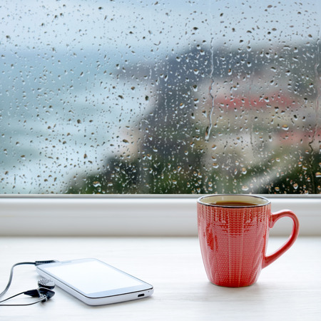 Cup of coffee, smartphone and headphones on a windowsill. In the background window with raindrops and cloudsの写真素材