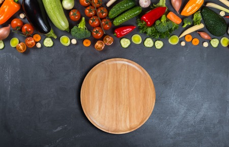 Different vegetables and empty wooden round plate on a black background for displaying productsの写真素材
